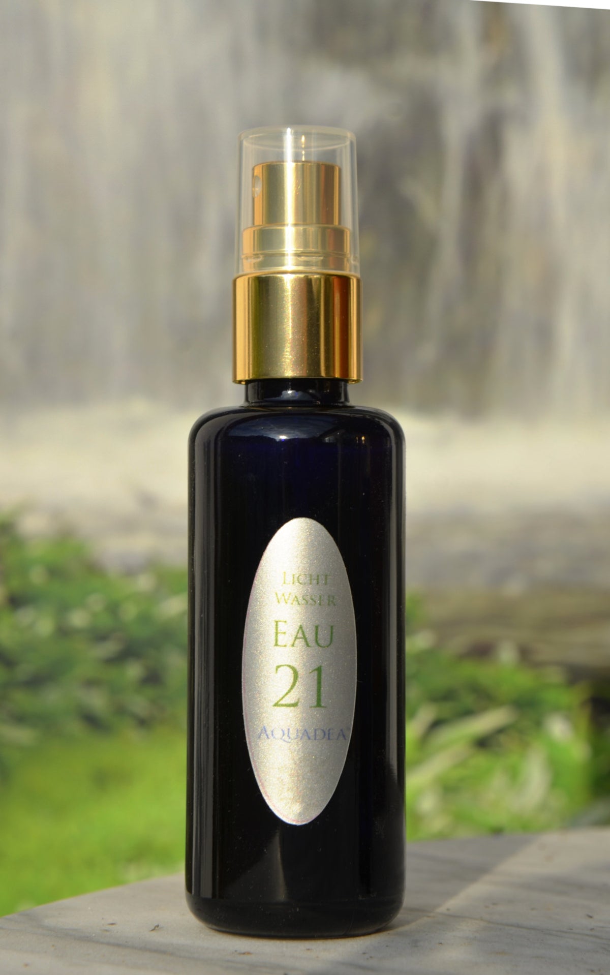 Eau 21 – Unio Mystica 21 Wasser in dunkler Glas-Sprühflasche mit goldenem Sprühkopf und silbernem Etikett „Licht Wasser Eau 21“, vor einem Wasserfall